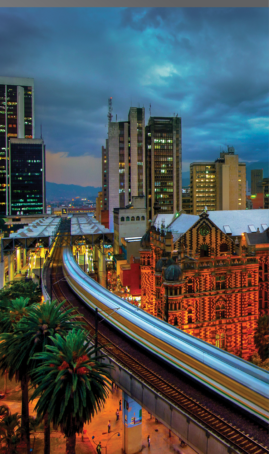 The elevated Medellin Metro is in motion as it rushes into the Parque Berrio Station in front of the illuminated Palace of Culture in Plaza Botero in Medellin, Colombia. The City of Eternal Spring is located in the Aburra Valley, a central region of the Andes Mountains in South America.