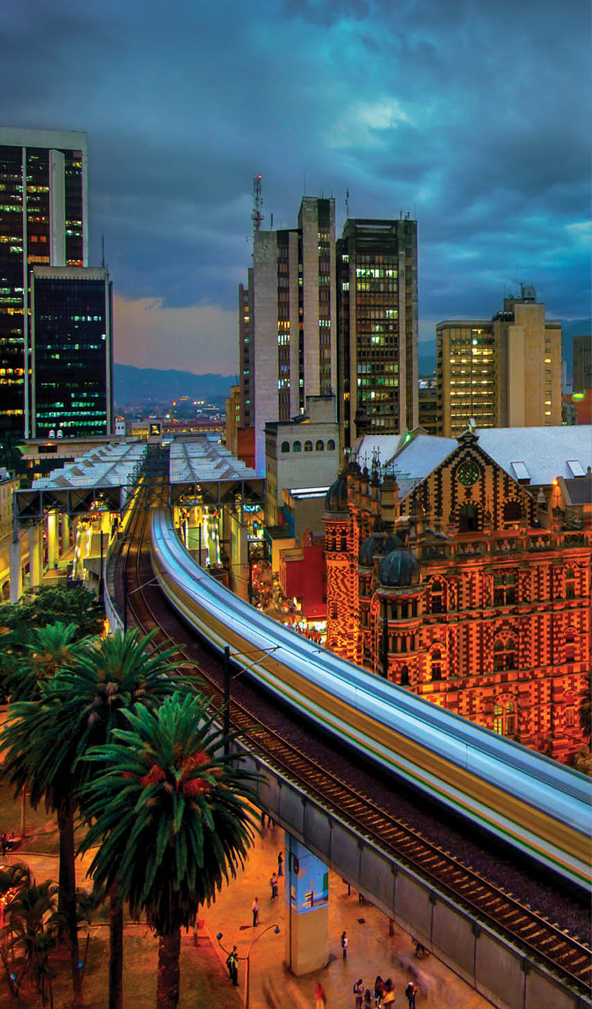 The elevated Medellin Metro is in motion as it rushes into the Parque Berrio Station in front of the illuminated Palace of Culture in Plaza Botero in Medellin, Colombia. The City of Eternal Spring is located in the Aburra Valley, a central region of the Andes Mountains in South America.