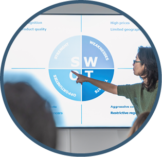 An adult Japanese woman confidently leads a meeting, pointing to a monitor displaying a SWOT analysis chart. Her attentive audience listens as she delves into strategic insights.