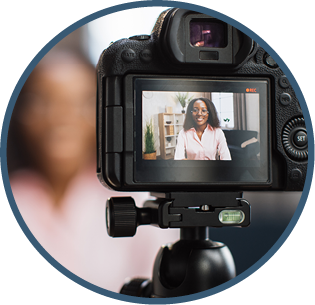 Smiling african woman talking and gesturing in front of modern video camera while sitting on blur background. Live streaming concept.