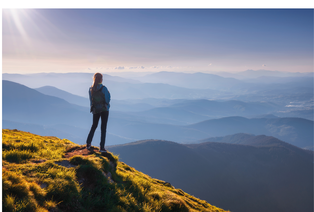 Girl with backpack on mountain peak with green grass looking at beautiful mountain valley in fog at sunset in summer. Landscape with sporty young woman, foggy hills, sky. Travel and tourism. Hiking