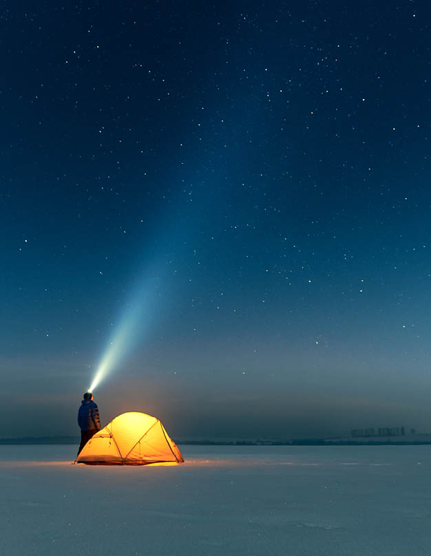 Tourist with flashlight near yellow tent lighted from the inside against the backdrop of incredible starry sky. Amazing night landscape. Tourists camp in snowy field. Travel concept