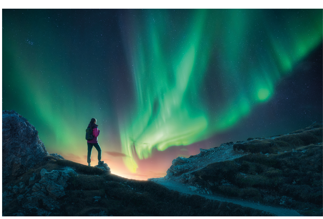 Northern lights and young woman on mountain peak at night. Aurora borealis and silhouette of alone girl on mountain trail. Landscape with polar lights. Starry sky with bright aurora. Travel background