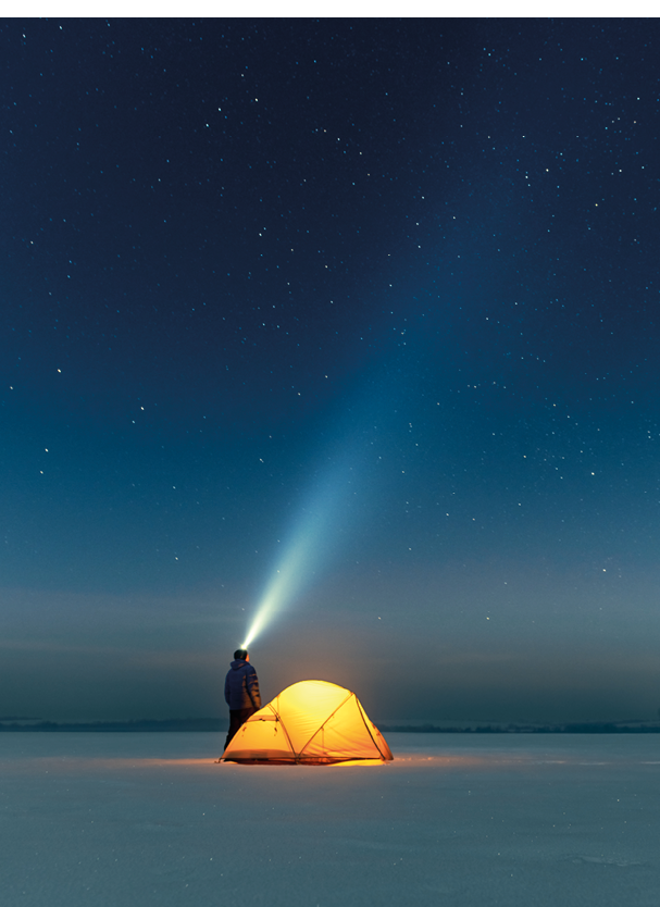 Tourist with flashlight near yellow tent lighted from the inside against the backdrop of incredible starry sky. Amazing night landscape. Tourists camp in snowy field. Travel concept